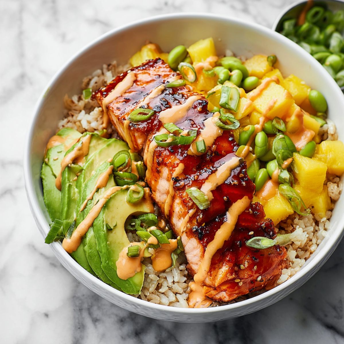 Salmon bowl with glazed teriyaki salmon fillet over brown rice topped with avocado slices edamame pineapple and spicy mayo sauce in white ceramic bowl