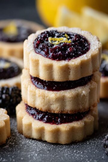 Shortbread cookies with jam centers stacked on dark slate showing buttery fluted edges and blackberry filling with fresh berries and lemon zest
