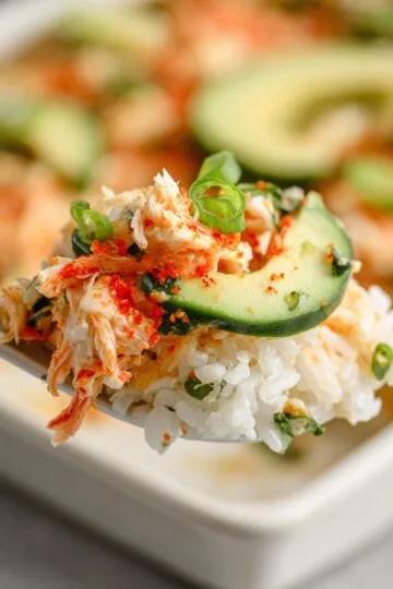 Sushi bake recipe showing spoonful of creamy crab mixture over seasoned rice topped with cucumber, avocado and nori with orange roe garnish on spoon