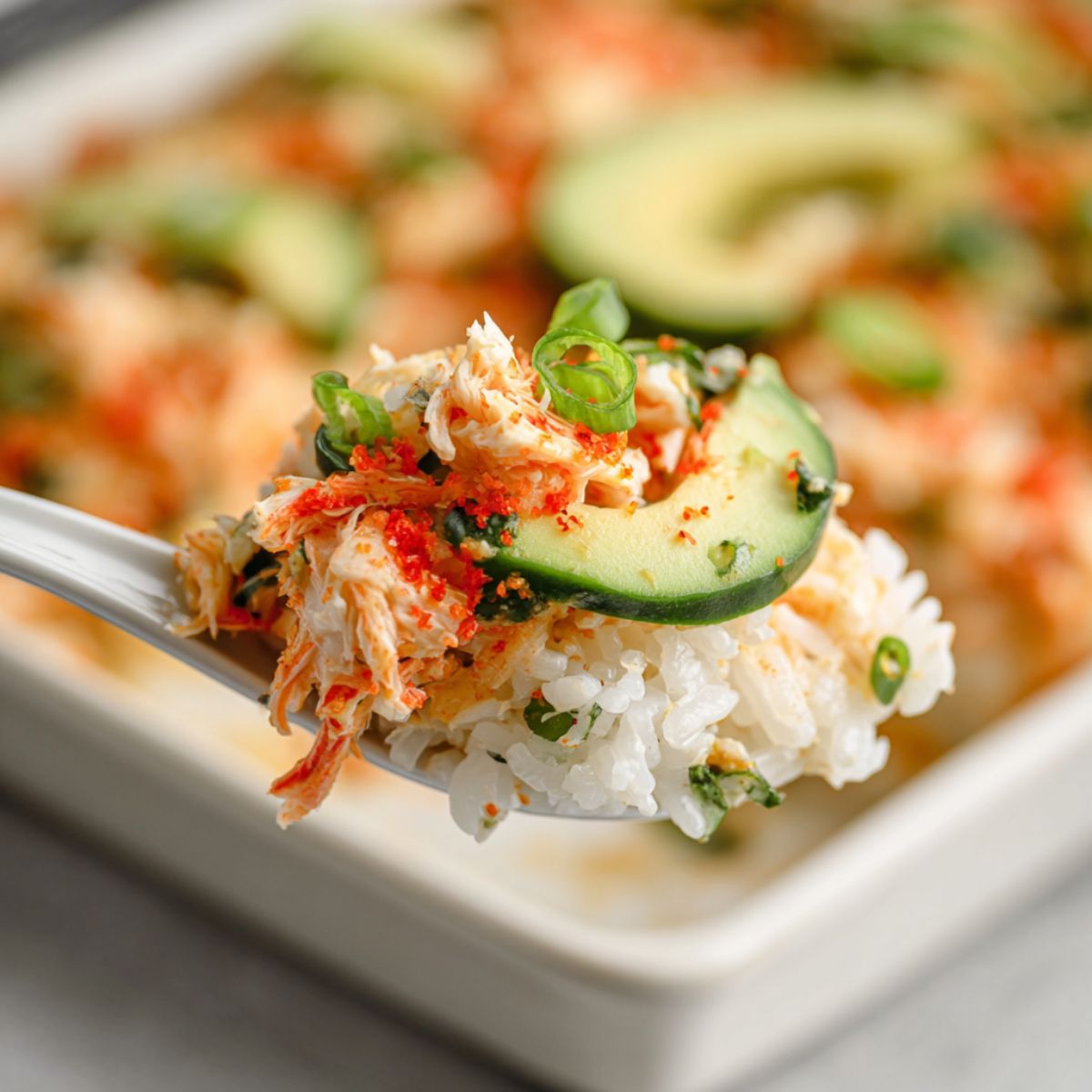 Sushi bake recipe showing spoonful of creamy crab mixture over seasoned rice topped with cucumber, avocado and nori with orange roe garnish on spoon