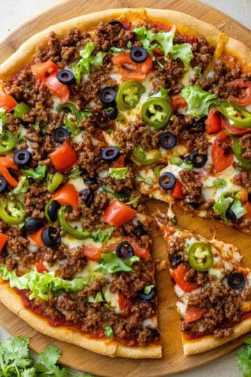 Taco pizza with seasoned ground beef black olives diced tomatoes jalapeños and lettuce on wooden cutting board sliced into triangles