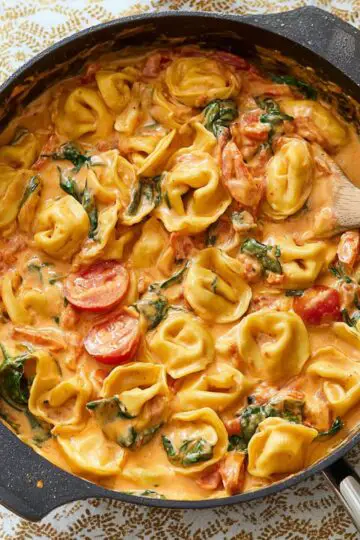 Tortellini in creamy garlic sauce with spinach and tomatoes cooking in cast iron skillet on wooden table