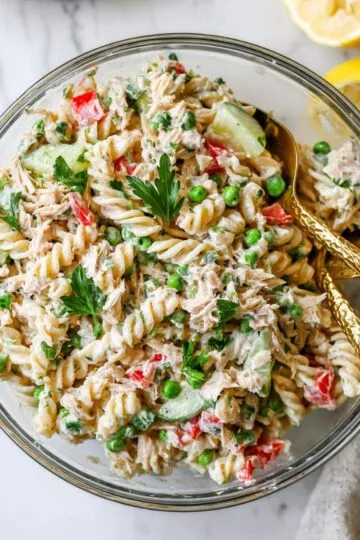 Tuna pasta salad in glass bowl with gold serving spoons showing rotini pasta mixed with tuna, peas, red peppers, cucumber and fresh parsley with lemon slices
