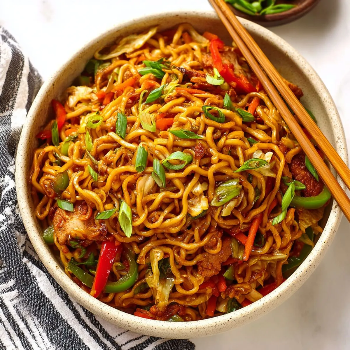 Asian noodles in ceramic bowl with chopsticks showing golden brown noodles mixed with red bell peppers green onions and savory sauce