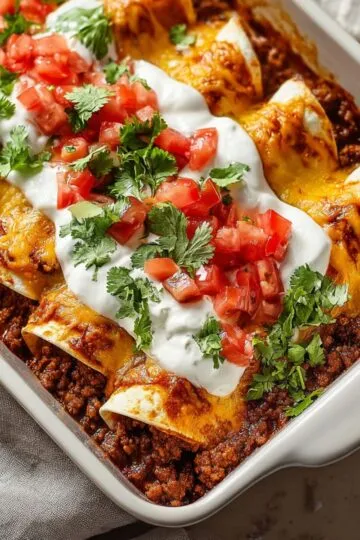 Beef enchiladas in white baking dish topped with melted cheese, sour cream, diced tomatoes and fresh cilantro showing golden tortillas filled with seasoned ground beef