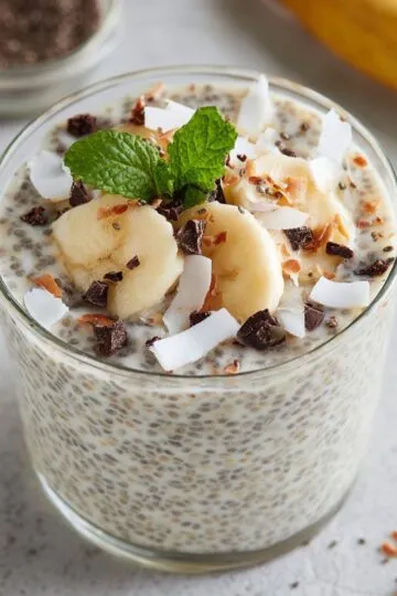 Chia seed pudding in glass jar topped with banana slices coconut flakes dark chocolate chunks and fresh mint garnish