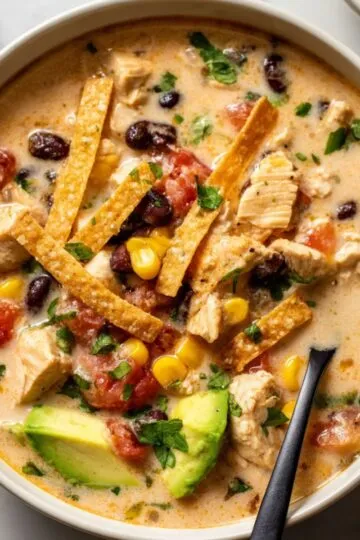Chicken taco soup in white bowl topped with crispy tortilla strips, diced avocado, black beans, corn and fresh cilantro showing creamy broth with tender chicken