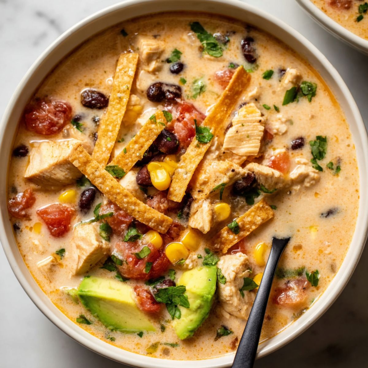 Chicken taco soup in white bowl topped with crispy tortilla strips, diced avocado, black beans, corn and fresh cilantro showing creamy broth with tender chicken