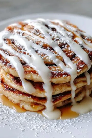 Cinnamon roll pancakes stack with white glaze drizzled on top, cinnamon swirl patterns visible, powdered sugar dusting on white plate