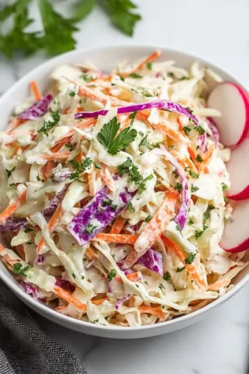 Coleslaw recipe showing fresh colorful mix of shredded green and purple cabbage with julienned carrots in creamy dressing garnished with fresh parsley and sliced radishes in white bowl