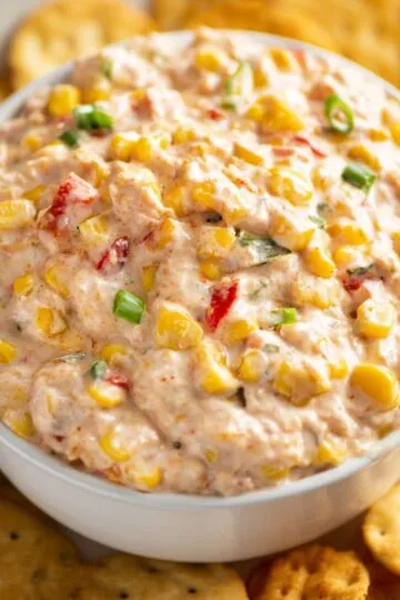 Corn dip with cream cheese in white bowl topped with golden corn kernels, red peppers and green onions surrounded by round crackers