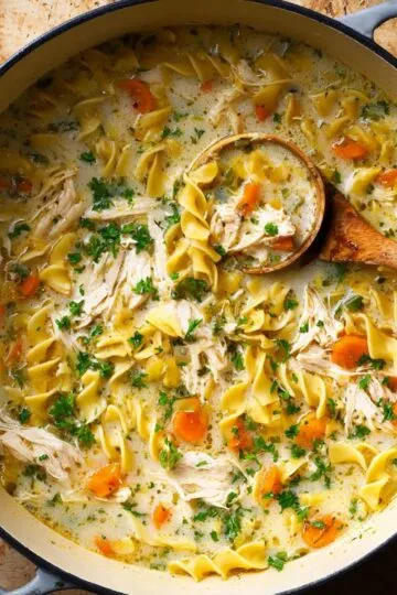 Creamy chicken noodle soup with shredded chicken, egg noodles, carrots and fresh parsley in one pot with ladle showing creamy broth texture