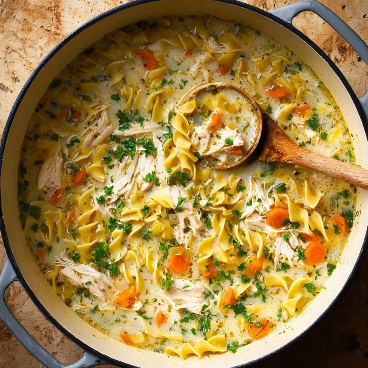 Creamy chicken noodle soup with shredded chicken, egg noodles, carrots and fresh parsley in one pot with ladle showing creamy broth texture