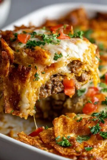Dorito casserole close-up showing melted cheese stretching from fork with ground beef filling and crushed Doritos in white baking dish