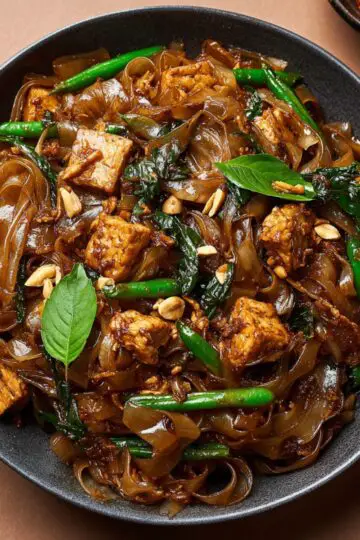 Drunken noodles close-up showing glossy wide rice noodles with tofu, fresh basil leaves, and green beans in black wok