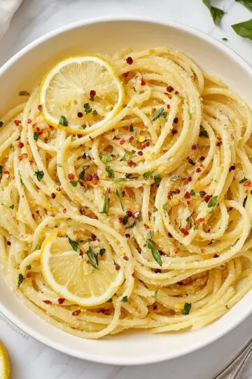 Garlic butter pasta in white bowl showing golden spaghetti coated in silky butter sauce with fresh parsley, red pepper flakes and lemon slices