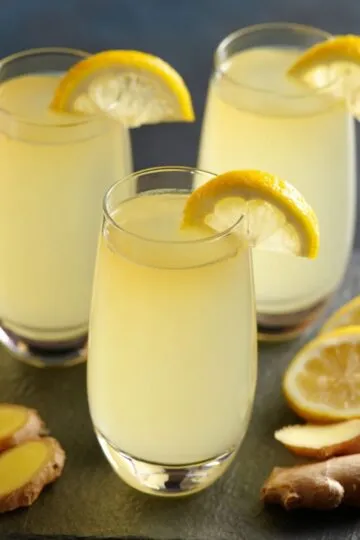 Ginger shot recipe in three glasses with pale yellow liquid, lemon wheel garnishes, and fresh ginger root on dark slate surface