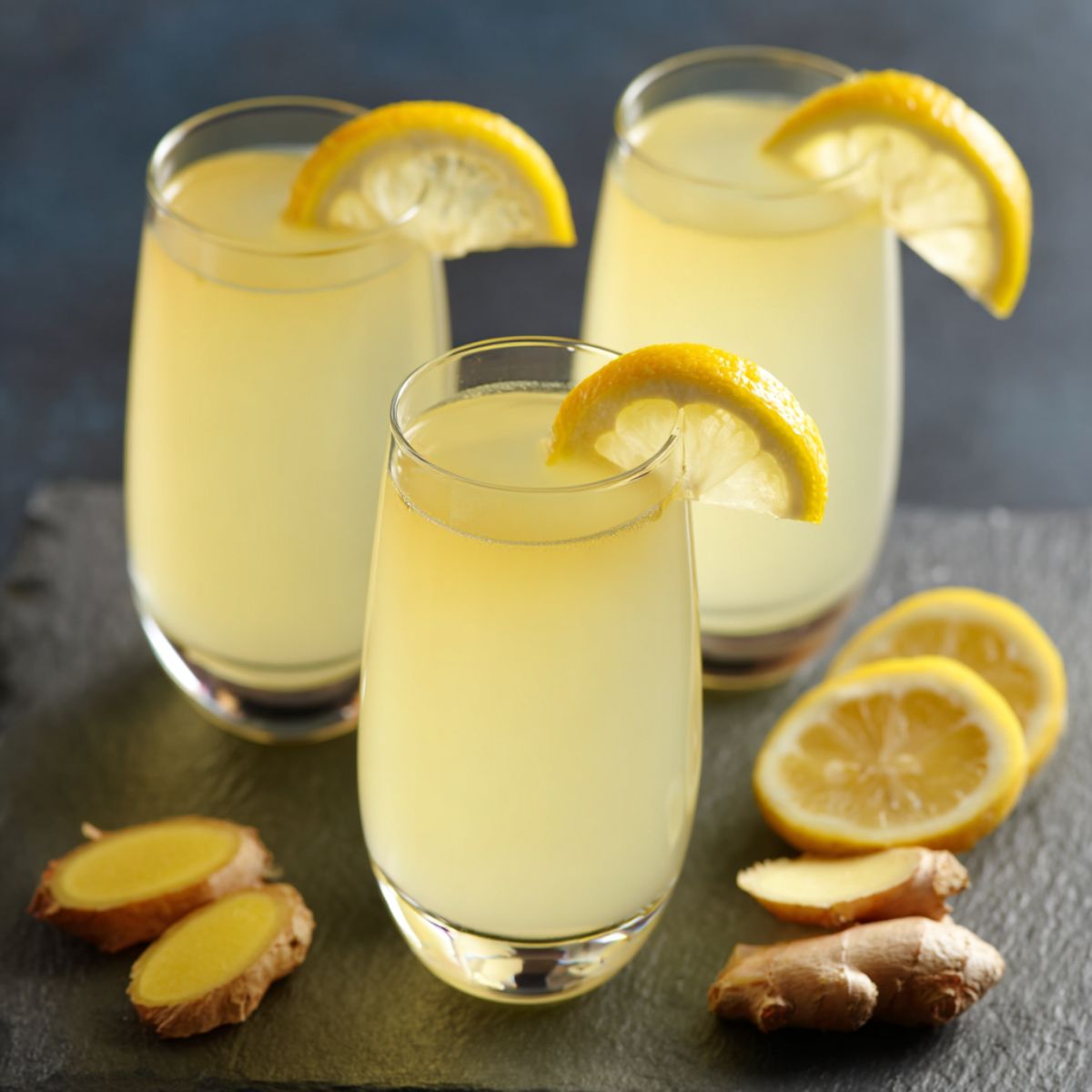 Ginger shot recipe in three glasses with pale yellow liquid, lemon wheel garnishes, and fresh ginger root on dark slate surface