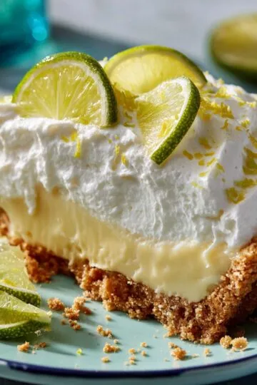 Key lime pie slice with creamy yellow filling, fluffy whipped cream topping, fresh lime wheel garnishes, and golden graham cracker crust on turquoise plate