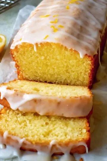 lemon cake glazed loaf with two slices cut, showing a moist, fluffy texture, garnished with fresh lemon zest and a lemon on a marble surface.
