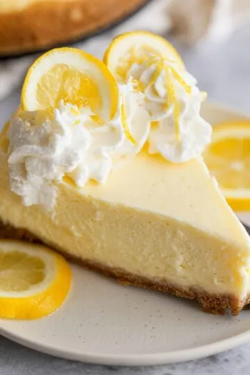 Lemon cheesecake slice with creamy yellow filling, whipped cream topping, fresh lemon slices garnish on white plate with graham cracker crust