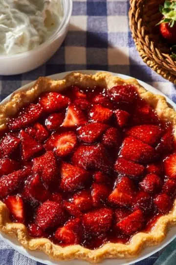 Strawberry pie recipe showing fresh glazed strawberries in golden flaky crust with whipped cream and fresh berries on checkered tablecloth