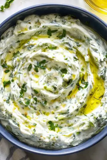 Tzatziki sauce in blue ceramic bowl with creamy white texture, fresh dill and herbs mixed throughout, drizzled with olive oil and surrounded by fresh herbs on marble surface