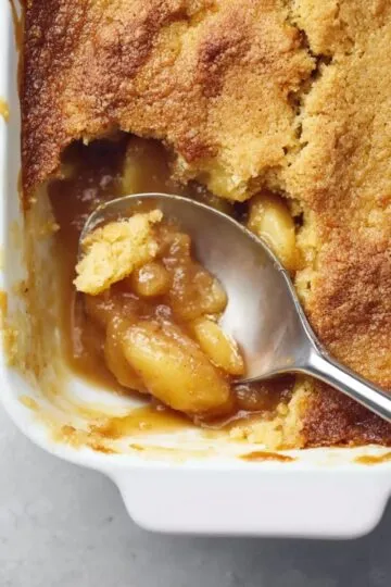 Apple dump cake with golden brown crispy topping, spoon scooping caramelized apple filling from white baking dish