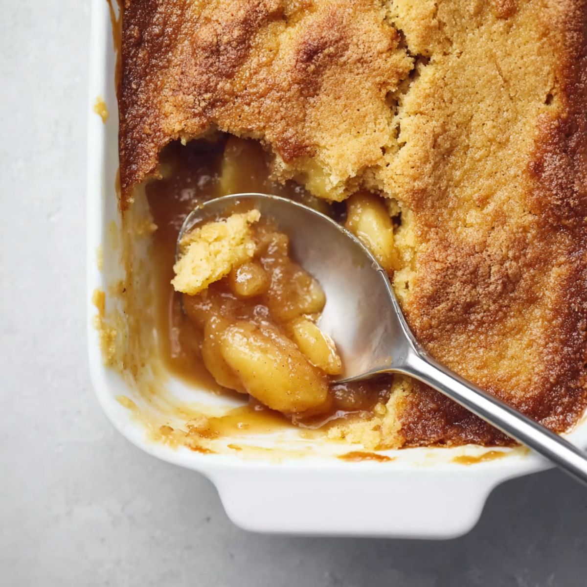 Apple dump cake with golden brown crispy topping, spoon scooping caramelized apple filling from white baking dish