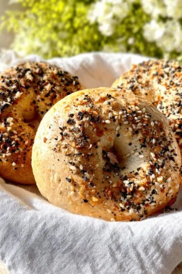 homemade bagel recipe A close-up shot of three homemade everything bagels covered generously with everything bagel seasoning, sitting in a white cloth basket. Soft sunlight is illuminating the bagels.