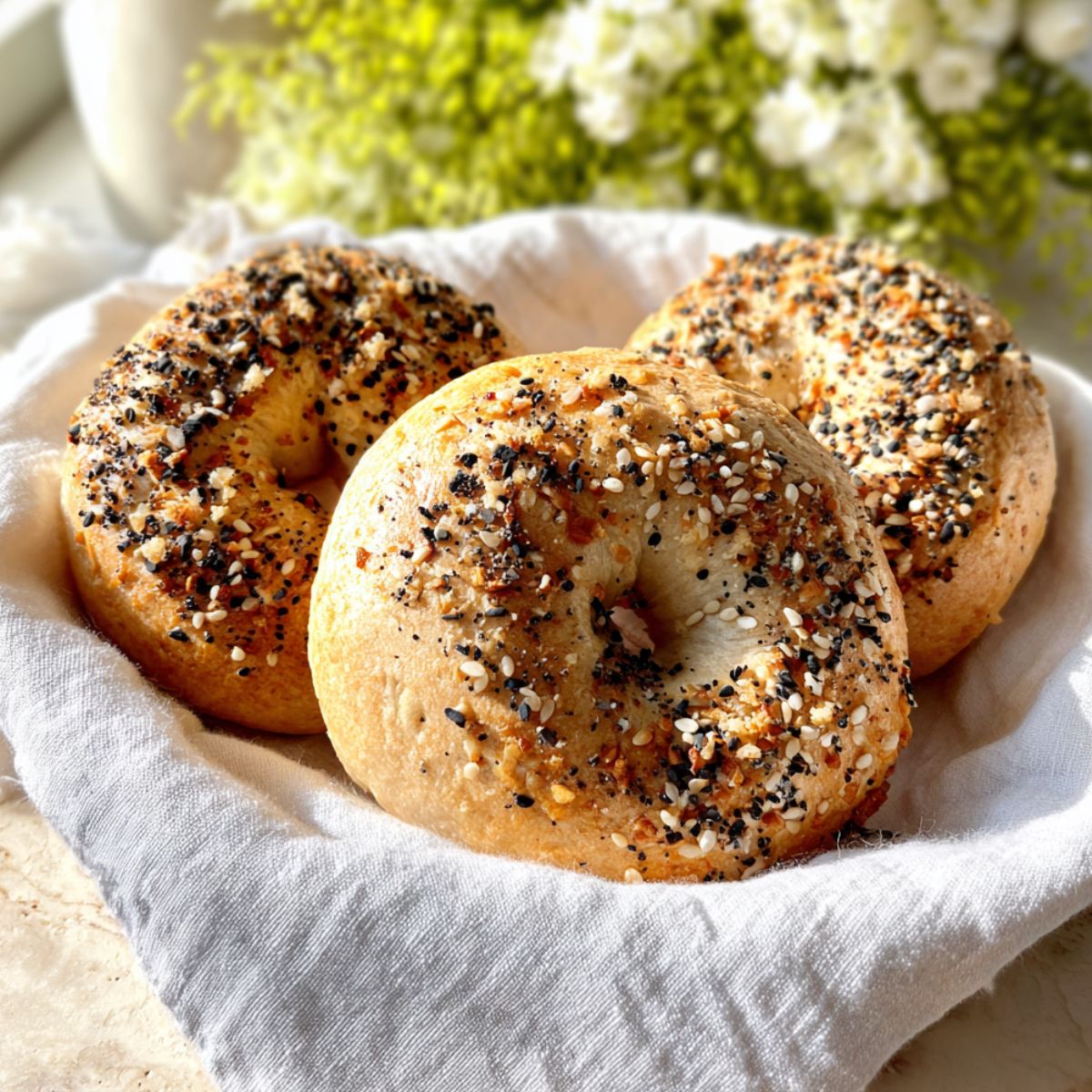 homemade bagel recipe A close-up shot of three homemade everything bagels covered generously with everything bagel seasoning, sitting in a white cloth basket. Soft sunlight is illuminating the bagels.