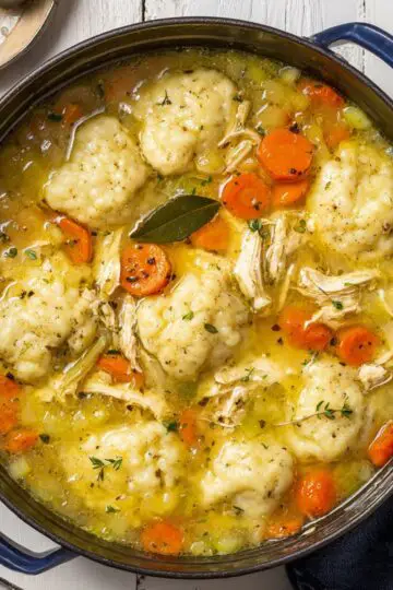 Chicken dumpling soup in blue pot showing fluffy homemade dumplings, shredded chicken, carrots and celery in golden creamy broth with herbs