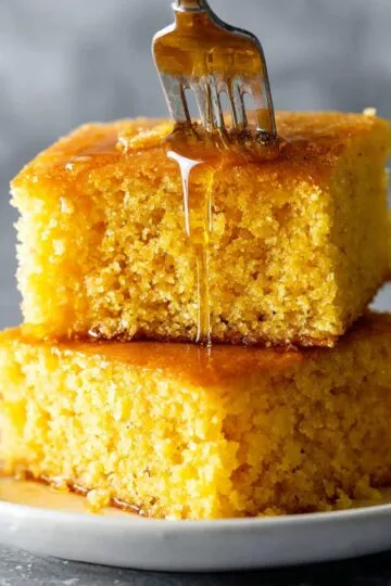 Cornbread recipe two golden squares stacked with honey being drizzled from fork showing moist texture on white plate