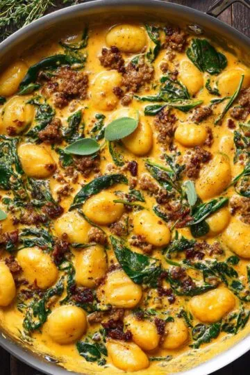Pumpkin gnocchi in creamy golden pumpkin sauce with crispy Italian sausage, wilted spinach and fresh sage leaves in large skillet on dark wooden surface with fresh thyme
