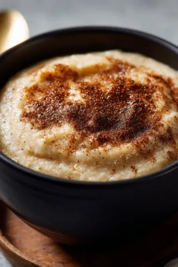 Crockpot applesauce smooth and creamy with cinnamon sprinkled on top in black bowl on wooden trivet with gold spoon