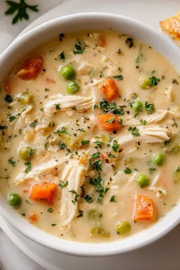 Chicken pot pie soup in white bowl with shredded chicken carrots peas and fresh herbs with flaky pastry on side