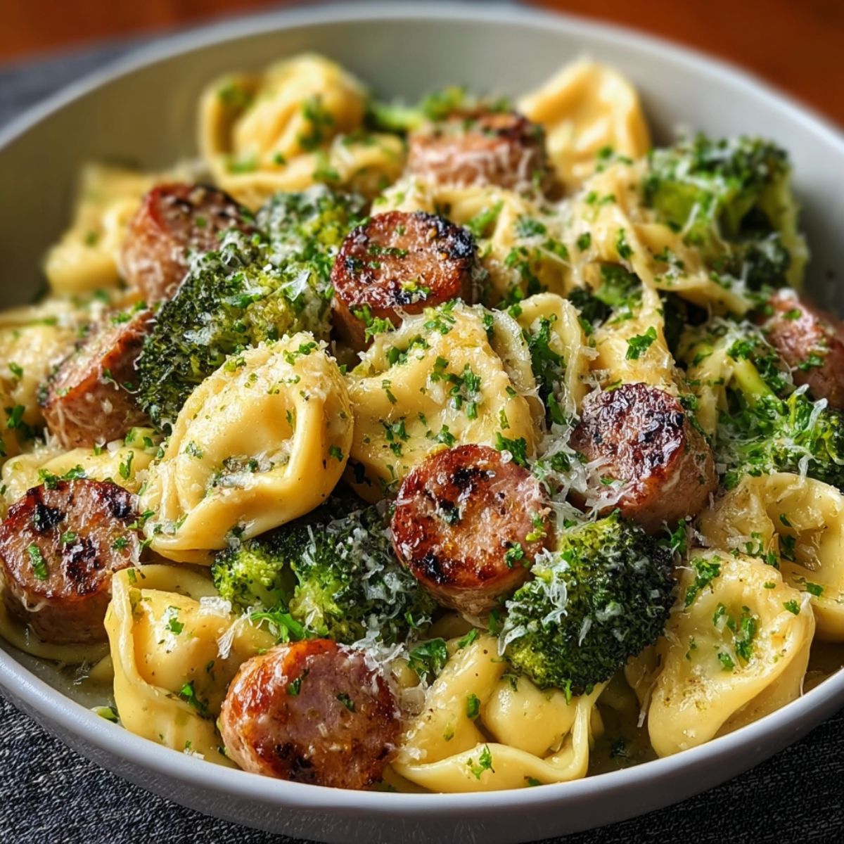 Garlic parmesan tortellini with sausage and broccoli in white bowl showing cheese-filled tortellini, crispy Italian sausage slices, tender broccoli florets topped with grated parmesan cheese and fresh parsley