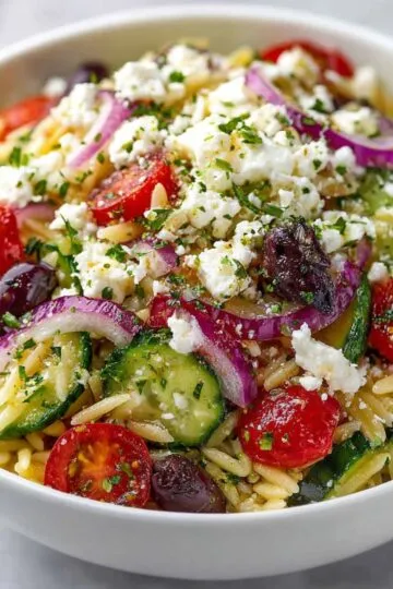 Mediterranean Orzo Pasta Salad in a white bowl topped with crumbled feta, fresh parsley, cherry tomatoes, cucumber slices, and purple onions
