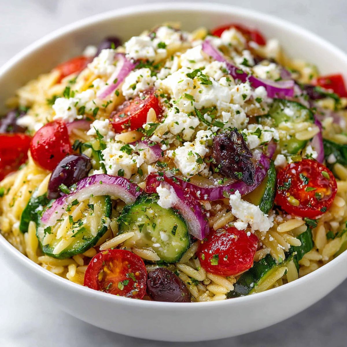 Mediterranean Orzo Pasta Salad in a white bowl topped with crumbled feta, fresh parsley, cherry tomatoes, cucumber slices, and purple onions