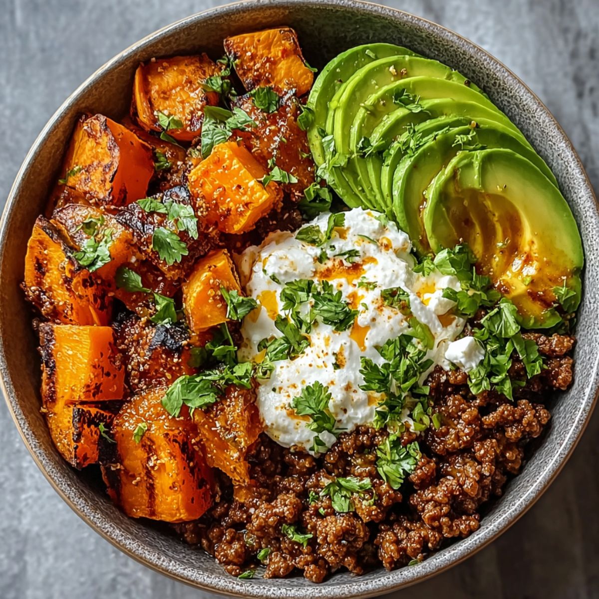 Ground beef hot honey bowl with caramelized sweet potato cubes, sliced avocado, seasoned ground beef, and creamy topping garnished with fresh cilantro in a rustic bowl