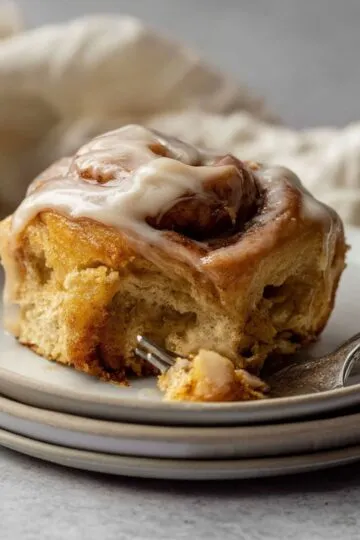Pumpkin cinnamon rolls with cream cheese frosting on white plate showing soft fluffy layers with cinnamon swirls and glaze dripping down sides with fork taking bite