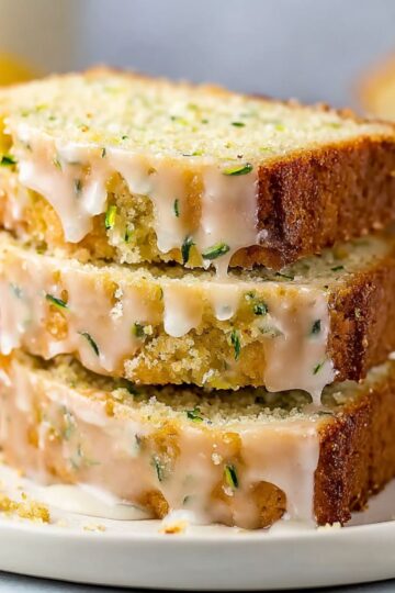 Lemon zucchini bread slices stacked with glossy lemon glaze dripping down golden edges and fresh lemon slices on white plate
