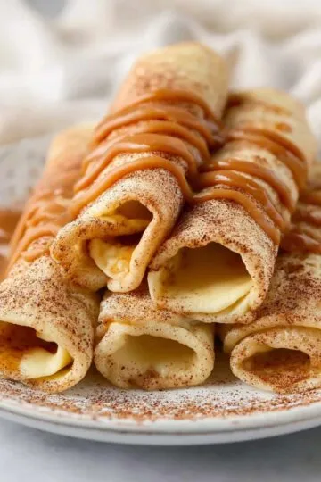 Crepe recipe featuring thin rolled French crepes dusted with cinnamon and drizzled with caramel sauce on a white plate