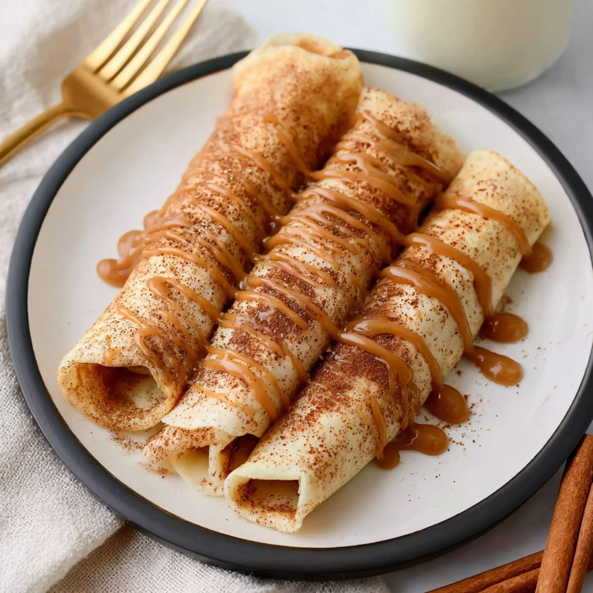 Crepe recipe showing rolled French crepes with cinnamon and caramel drizzle on a black-rimmed white plate with gold fork