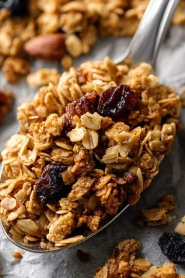 Granola recipe featuring golden crunchy clusters with oats, nuts, and dried cranberries on a silver spoon