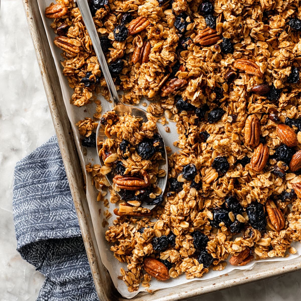 Granola recipe on a baking sheet with golden oat clusters, pecans, almonds, and dried blueberries with a silver spoon