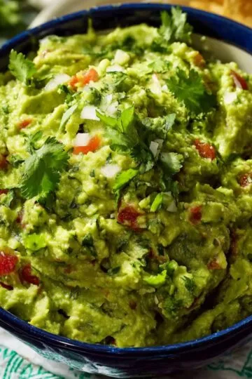 Guacamole recipe in blue bowl topped with cilantro, red onion, and tomatoes, chunky creamy texture, spoon ready to serve
