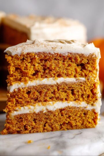 Pumpkin Cake slice on a marble coaster with the whole cake and orange pumpkins in the background.