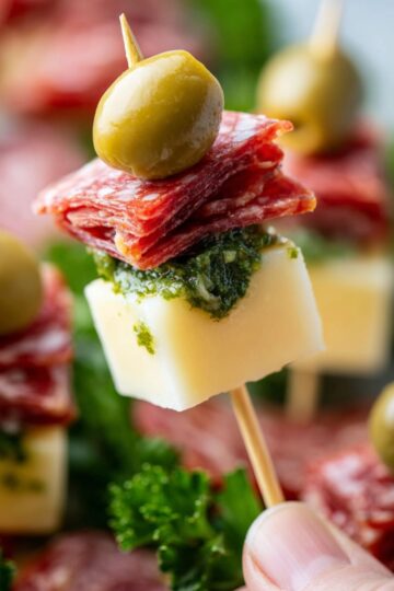 Hand holding a single salami and cheese skewer topped with pesto and olive.