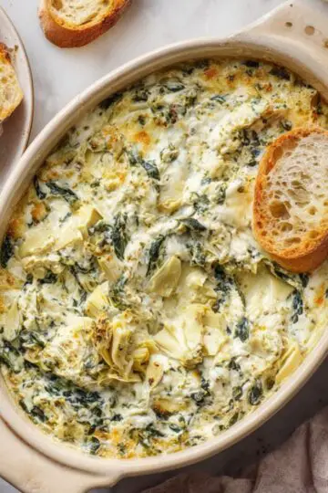 Golden brown baked spinach artichoke dip in oval dish with baguette